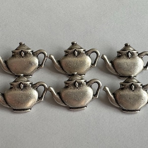 Puede incluir: Seis pequeños dijes de tetera de color plateado, dispuestos en dos filas de tres. Cada dije tiene un diseño detallado, que incluye una boquilla, un asa y una tapa. Las teteras están sobre un fondo gris claro.