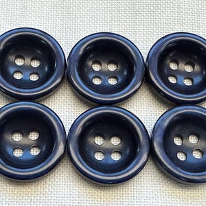 Puede incluir: Un conjunto de seis botones redondos de color azul marino oscuro dispuestos en dos filas de tres. Cada botón presenta un centro liso ligeramente hundido con cuatro agujeros para fijación, con un acabado brillante y un borde grueso.
