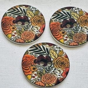 May include: Three large, round buttons with a floral design. The buttons feature a white background with a pattern of orange, yellow, and burgundy flowers and green leaves. Each button has two holes for sewing.