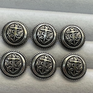 Könnte beinhalten: Sechs silberfarbene Knöpfe mit Ankerdesign. Jeder Knopf zeigt ein detailliertes Ankeremblem in einem kreisförmigen Rahmen, umgeben von einem seilartigen Rand. Die Knöpfe sind in einem ordentlichen Muster angeordnet.