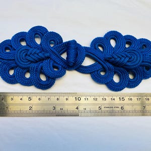 Op de afbeelding: Een paar blauwe Chinese knoop-sluitingen, ook wel bekend als knopen of sluitingen, met een decoratief ontwerp. De sluitingen zijn ongeveer 15 cm lang.