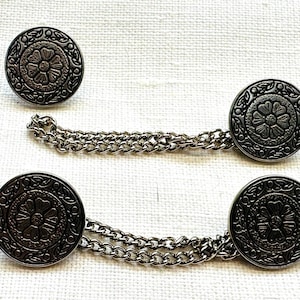 May include: Set of three silver-toned metal buttons with a floral design. Two buttons are connected by a chain. The buttons have a detailed, embossed pattern and are round in shape. The background is a white, textured fabric.