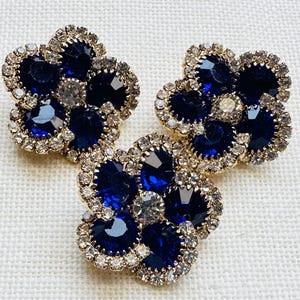 May include: Three flower-shaped brooches or pins featuring deep blue faceted crystals arranged as petals around a central clear crystal. Each piece is outlined with a border of small, sparkling clear rhinestones set in a gold-toned metal frame.