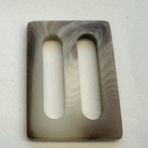 May include: A rectangular, off-white plastic buckle with two oval-shaped openings. The buckle has a swirling, marbled pattern in shades of brown and grey.