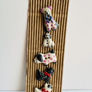 May include: A collection of five dog-shaped buttons in various colors and patterns. The buttons are attached to a corrugated cardboard backing. The dogs have different designs, including spots, bows, and contrasting colors.
