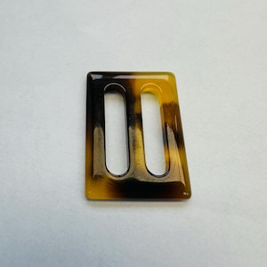 Peut inclure: Boucle rectangulaire avec deux ouvertures allongées. Le motif est en écaille de tortue, avec des nuances de marron et de jaune. Les bords sont lisses et la forme générale est légèrement trapézoïdale.