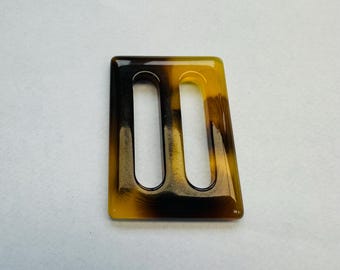 Puede incluir: Hebilla rectangular con dos aberturas alargadas. La hebilla presenta un patrón de carey con tonos marrones y amarillos. Los bordes son lisos y la forma general es ligeramente trapezoidal.