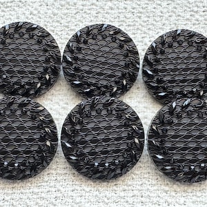 May include: Six black, circular buttons with a decorative design. Each button has a textured, wave-like pattern in the centre, encircled by a leaf-like border and small, dark embellishments. The buttons are arranged on a light-coloured surface.