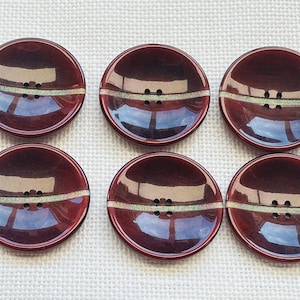 May include: A set of six round, dark burgundy buttons with a glossy finish. Each button features a concave centre and a horizontal, textured light green band running across the middle, with four small holes centred for attachment purposes.