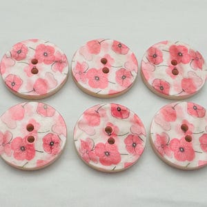Könnte beinhalten: Sechs runde Holzknöpfe mit floralem Design. Die Knöpfe haben einen hellen Holzrand und eine weiße Oberfläche, die mit rosa Mohnblumen verziert ist. Jeder Knopf hat drei Löcher zum Nähen.