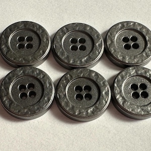 Puede incluir: Seis botones redondos de color gris oscuro con cuatro agujeros cada uno. Los botones tienen un aspecto de metal martillado texturizado y un borde ligeramente elevado. Adecuados para proyectos de costura y manualidades.