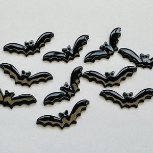 May include: Ten black bat-shaped embellishments with a glossy finish. Each bat features a detailed wing design with a lighter, striped pattern. These decorative bats are suitable for Halloween crafts and decorations.