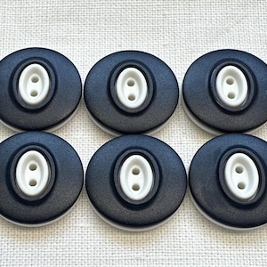 May include: A set of six circular buttons, each featuring a matte black outer ring and a raised, oval-shaped white center. Each white center contains two vertical holes for attachment. The buttons are arranged in two rows of three on a textured surface.