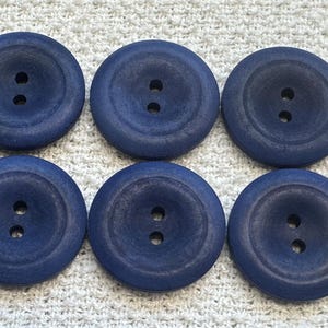 May include: Six navy blue wooden buttons with two holes each. The buttons are round with a slightly recessed center and a smooth, matte finish. They are arranged in two rows of three on a textured white surface.
