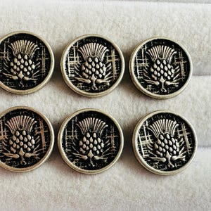 May include: Six silver-toned metal buttons with a thistle design. Each button features a detailed thistle emblem within a circular frame. The buttons are arranged in two rows of three, showcasing their decorative appeal. Ideal for sewing projects or adding a vintage touch.