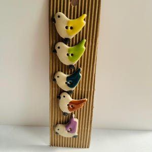 May include: Five ceramic bird-shaped buttons in yellow, green, teal, orange, and purple, attached to a brown corrugated cardboard backing. Each bird has a white body and black eyes. Ideal for craft projects.