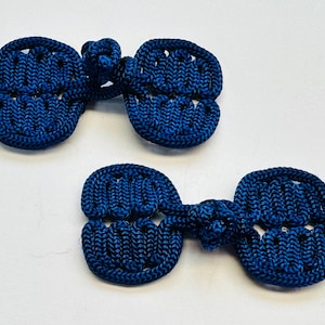 May include: Two navy blue frog closures, a traditional fastening method. Each closure features a knotted loop and two symmetrical, oval-shaped components with a woven texture. The closures are displayed against a white background.