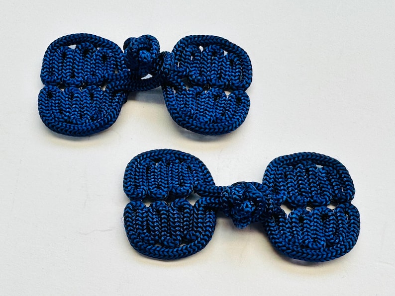 May include: Two navy blue frog closures, a traditional fastening method. Each closure features a knotted loop and two symmetrical, oval-shaped components with a woven texture. The closures are displayed against a white background.