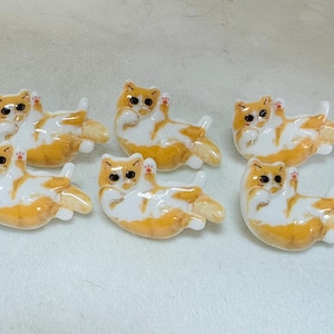 May include: Six ceramic cat figurines, each with a white body and orange markings. The cats are in various poses, with some lying down and others sitting up. They have black eyes and pink noses.