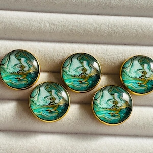 May include: Five circular buttons with a gold edge. Each button has a swirling design in teal, white, and gold, like an abstract landscape. They are presented on a soft, light-coloured surface.