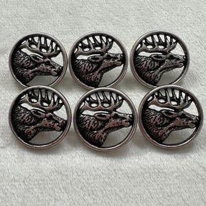 May include: A set of six circular metal buttons, each featuring a detailed, raised relief design of a stag head with antlers. The buttons have a dark, oxidised silver finish with recessed areas that create contrast against the metallic surface.