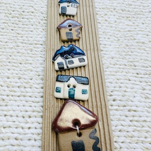 May include: Five handmade ceramic buttons shaped like houses, arranged vertically on a corrugated cardboard strip. Each button features unique hand-painted details, including blue, brown, and metallic gold roofs, with white or tan walls and black accents.