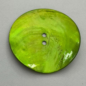 May include: A large, round, lime green button with two small holes. The button has a glossy, iridescent finish, reflecting light. The button is a sewing or craft supply.