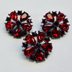 Peut inclure: Trois bijoux circulaires dotés de montures en métal sombre et de cristaux rouges facettés disposés en motif floral. Chaque pièce contient plusieurs pierres rouges en forme de poire et rondes regroupées pour former un motif de fleur symétrique.