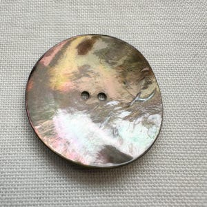 May include: A round, two-hole button featuring a polished, iridescent surface with swirling shades of brown, tan, and subtle hints of pink and green. The button has a smooth, reflective finish that catches the light, displaying natural variations in colour across its circular face.
