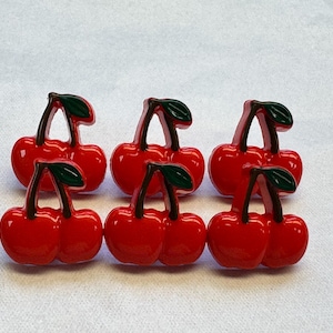 Puede incluir: Seis adornos brillantes en forma de cereza roja con hojas verdes oscuras y tallos marrones. Las cerezas están dispuestas en dos filas de tres, sobre un fondo blanco. Ideales para manualidades o bisutería.