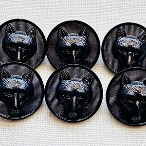 Puede incluir: Seis botones negros con un diseño detallado de cabeza de lobo. Cada botón presenta una cara de lobo en relieve dentro de un marco circular con un fondo texturizado. Los botones están hechos de un material oscuro y brillante.