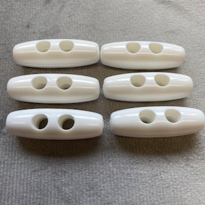 May include: Six white plastic toggle clasps, each with two holes. The oval-shaped clasps are arranged on a grey fabric surface. These are commonly used for closures on jackets, bags, and other apparel.
