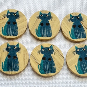 May include: Six round wooden buttons, each featuring a printed illustration of a blue cat with long whiskers and two holes for attachment. The natural wood grain is visible on the surface of every button, showcasing a light tan colour.
