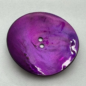Peut inclure: Gros bouton rond avec une finition irisée violette vibrante. Le bouton comporte deux petits trous près du centre pour la couture. Les bords du bouton sont d'un violet plus foncé, créant un contraste subtil.