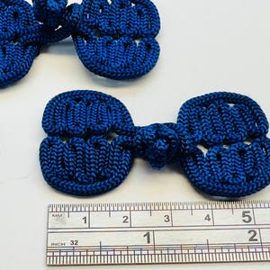 May include: Close-up of several navy blue frog closures. Each closure is approximately 2 inches long. The closures are made of a woven material and are used for decorative purposes on clothing or accessories.
