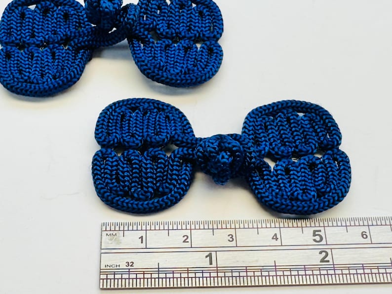 May include: Close-up of several navy blue frog closures. Each closure is approximately 2 inches long. The closures are made of a woven material and are used for decorative purposes on clothing or accessories.