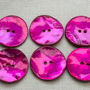 May include: A set of six round, vibrant magenta buttons, each featuring a smooth, iridescent surface with a natural shell-like texture and two centered holes for attachment, arranged in two rows of three on a neutral fabric surface.