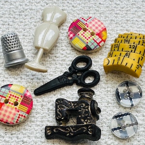 May include: A selection of sewing-related decorative items. The image shows a silver thimble, a cream-coloured dress form, black scissors, a yellow measuring tape, and several buttons with patchwork patterns. A small black sewing machine is also visible.