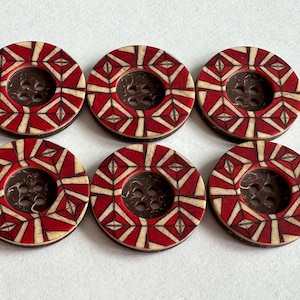 Op de afbeelding: Zes ronde decoratieve knopen. Elke knoop heeft een rood en crèmekleurig geometrisch patroon langs de rand, met een donkerbruin midden. De knopen zijn in twee rijen van drie gerangschikt.