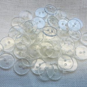 May include: A collection of numerous round, clear plastic buttons, each featuring two small holes in the centre for attachment. These transparent, circular fasteners are arranged in a loose pile, showcasing their smooth, glossy surface and simple design.