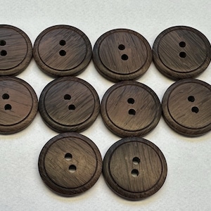May include: Ten dark brown wooden buttons, each with two holes. The buttons have a raised outer rim and a visible wood grain. These buttons are suitable for sewing and craft projects. Each button is approximately 2.5 cm in diameter.