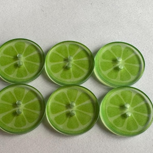 Puede incluir: Seis botones redondos y transparentes con un diseño de rodaja de lima. Cada botón tiene un centro verde claro con un patrón verde más oscuro, que se asemeja a una lima. Los botones tienen dos agujeros para coser y están colocados sobre una superficie blanca.