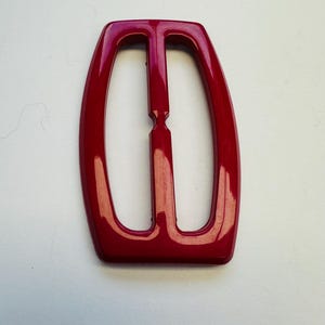 May include: A glossy, red buckle with a rectangular shape and a central bar. The buckle has rounded edges and a smooth surface, suitable for belts or other accessories. The design is simple and modern.