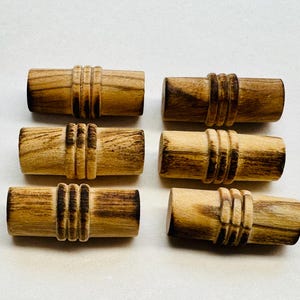 May include: Six wooden toggle clasps with a natural wood grain. Each clasp is cylindrical with three carved, grooved bands. The clasps are arranged on a white surface, showcasing their warm, earthy tones.