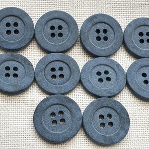 May include: A set of ten round, dark gray buttons, each featuring a recessed center with four evenly spaced holes. The buttons have a matte, textured surface and are arranged in a grid pattern, showcasing a consistent circular shape and uniform design.