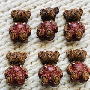 May include: Six small brown teddy bear buttons arranged in two rows. Each bear features a textured brown fur finish and is wearing shiny dark red overalls with white painted buttons on the straps. The bears are depicted in a seated position.