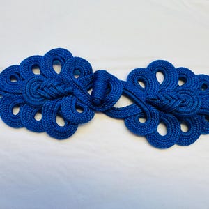 Op de afbeelding: Een paar blauwe stoffen Chinese knoop-sluitingen, ook wel bekend als een kikkersluiting, met een decoratief ontwerp.
