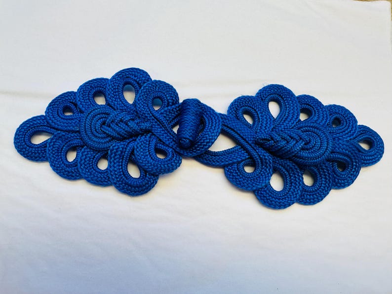 Op de afbeelding: Een paar blauwe stoffen Chinese knoop-sluitingen, ook wel bekend als een kikkersluiting, met een decoratief ontwerp.