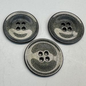 May include: Three metallic gray buttons with four holes each. The buttons have a shiny, speckled finish and a slightly raised edge. These buttons are suitable for sewing projects and garment construction.