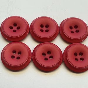 May include: Six red buttons with four holes each are arranged in a close-up shot. The buttons have a slightly raised centre and a rounded edge. The buttons are a solid, matte red colour.